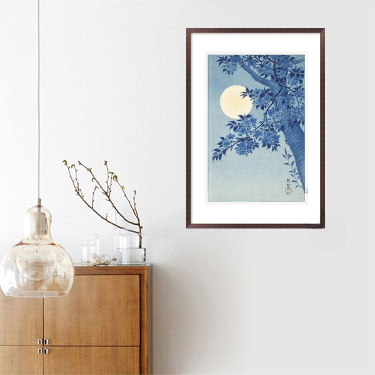 Framed artwork of a blue floral design with a moon on a white wall above a wooden cabinet.