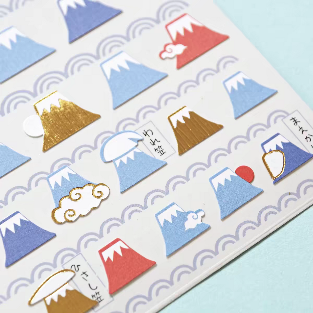 A close-up of a sheet of decorative stickers featuring stylized Mount Fuji illustrations in blue, red, white, and gold foil, arranged on a white background with a light blue surface visible at the edges.