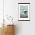 Framed artwork of birds flying over water on a white wall above a wooden cabinet.