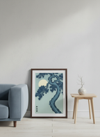 Framed artwork of a tree in a room with a gray armchair and wooden side table.